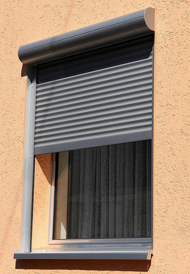 Volet roulant gris foncé recouvrant partiellement une fenêtre sur un bâtiment orange clair.