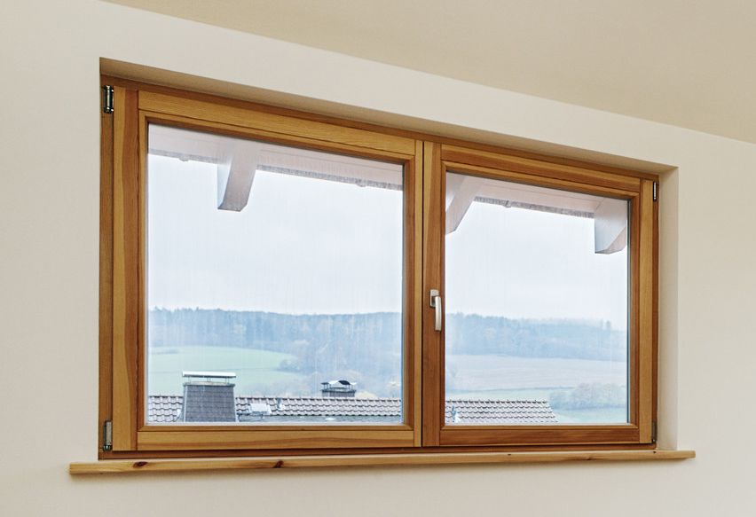 Fenêtre à cadre en bois dans une pièce aux murs blancs, donnant sur un paysage rural avec des arbres et un ciel nuageux.