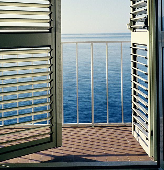 Des volets ouverts encadrent une vue sur l'océan bleu depuis un petit balcon avec une balustrade blanche.