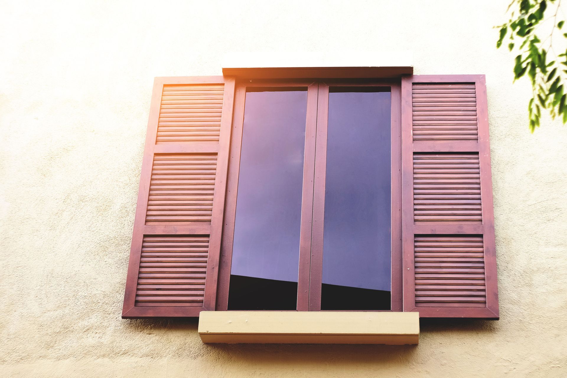 Fenêtre en bois avec volets ouverts sur un mur en stuc beige, baigné de soleil.