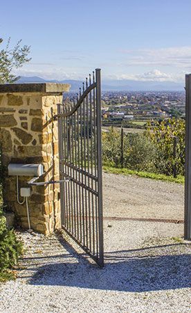 Portail en fer forgé ouvert dans un mur de pierre, allée de gravier menant à la ville au loin, journée ensoleillée.