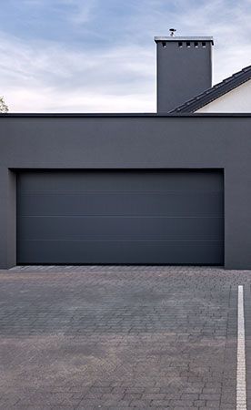 Porte de garage grise sur un bâtiment gris avec une allée en briques et une cheminée, contre un ciel nuageux.