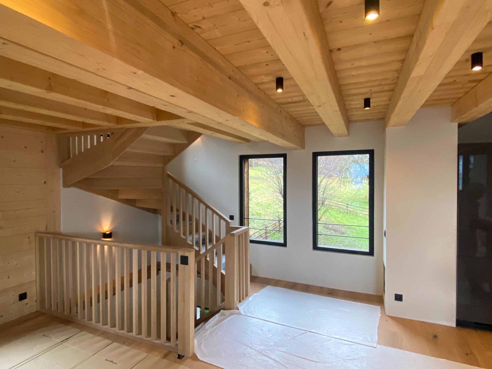 Intérieur d’une maison moderne en bois avec poutres apparentes au plafond. Escalier en bois menant à un étage supérieur. Trois grandes fenêtres laissent entrer la lumière naturelle et offrent une vue sur la verdure. Sol protégé par des bâches, suggérant des travaux en cours. Éclairage par spots encastrés au plafond.