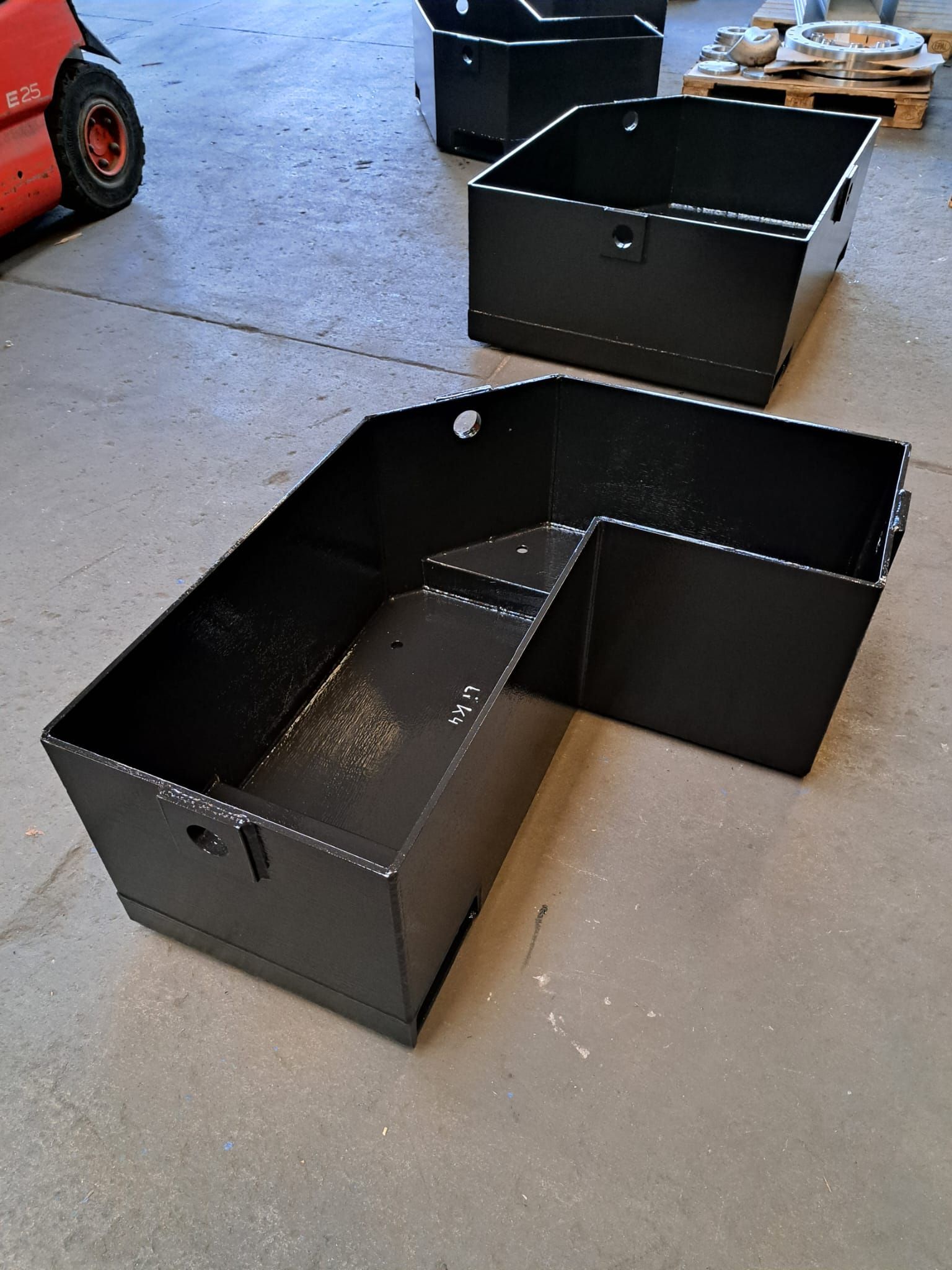 Drei schwarze Kästen stehen auf einem Betonboden neben einem Gabelstapler.