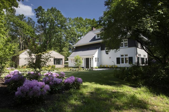 Wit huis met donker dak en luiken, omgeven door bomen en een tuin met paarse bloemen.