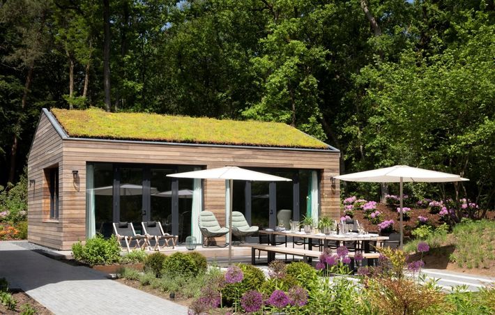 Houten blokhut met groen dak, grote ramen en een eethoek buiten. Omgeven door een weelderige tuin en bomen.