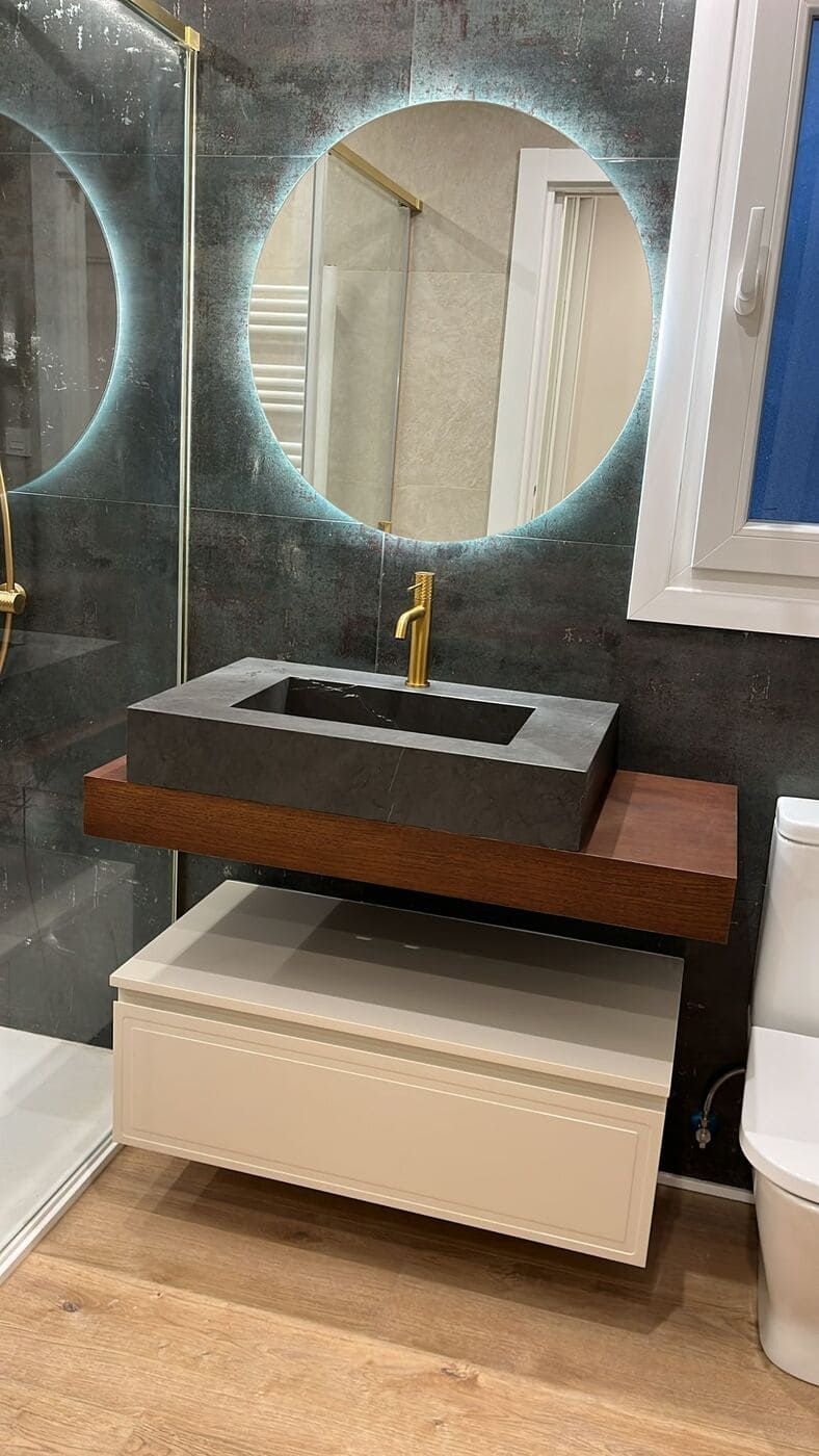 Baño con tocador flotante, espejo redondo iluminado, lavabo gris oscuro, grifería dorada y mueble blanco.