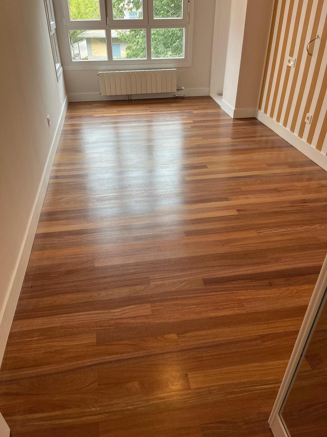 Piso de madera en una habitación vacía con una ventana, molduras blancas y una pared estampada.