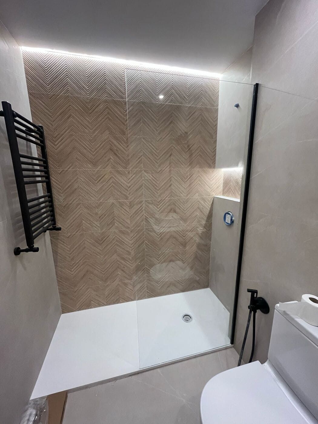 Ducha con puerta de vidrio, detalles en azulejos de espiga beige, toallero negro e inodoro blanco.
