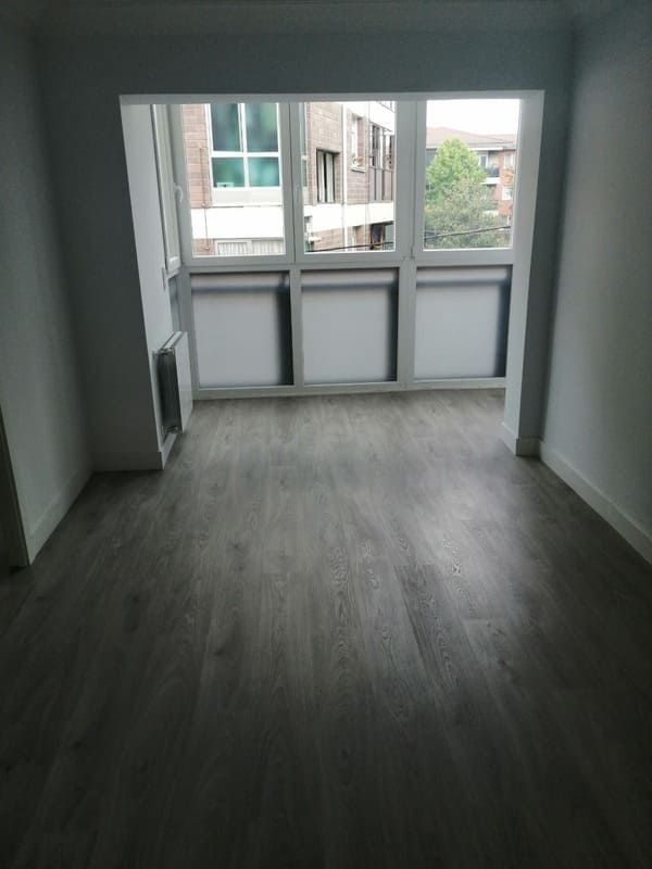 Habitación vacía con piso de apariencia de madera gris, ventana grande de varios paneles y paredes blancas.