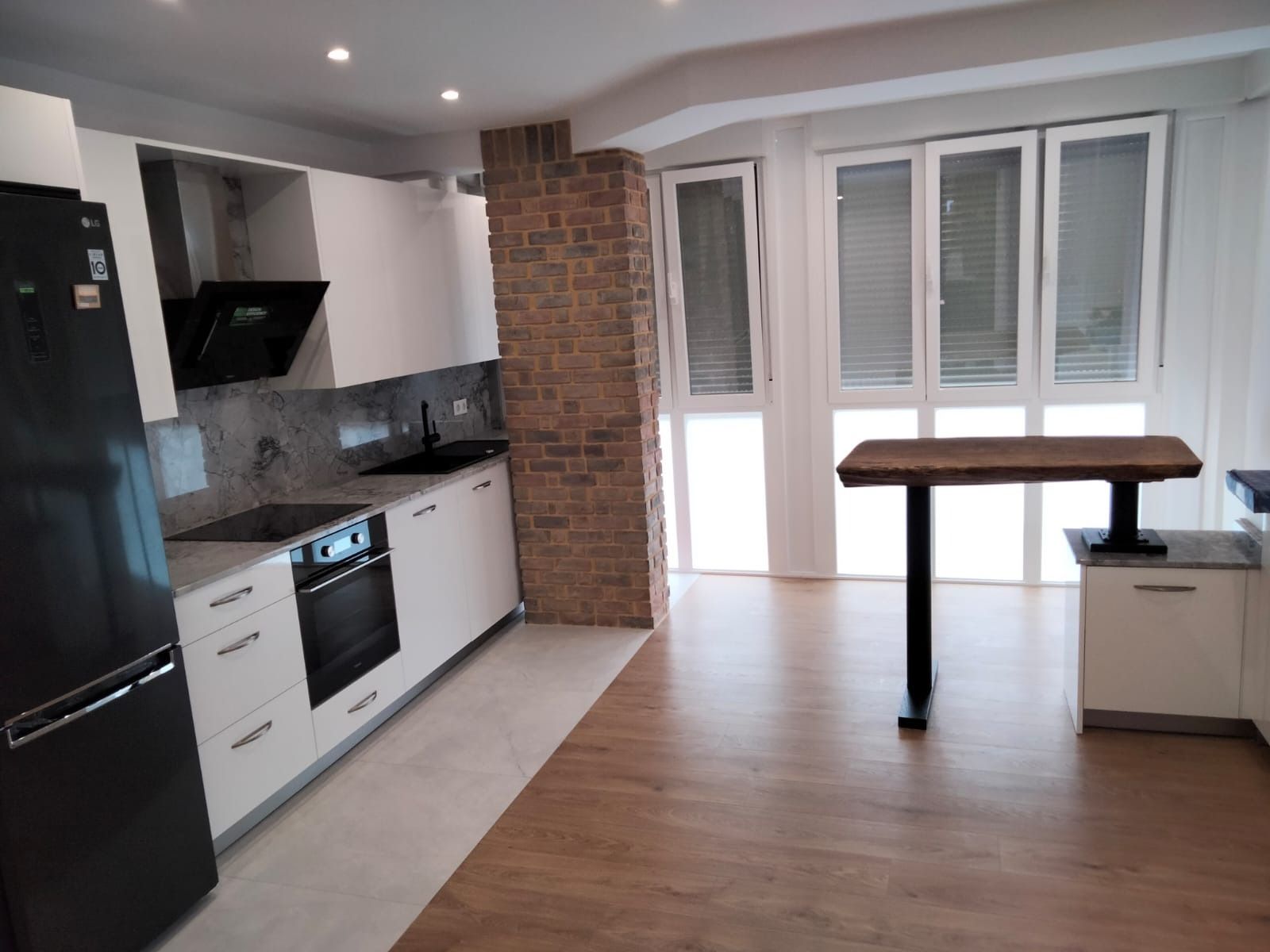 Cocina moderna con gabinetes blancos, electrodomésticos oscuros, columna de ladrillo y una pequeña mesa de madera.