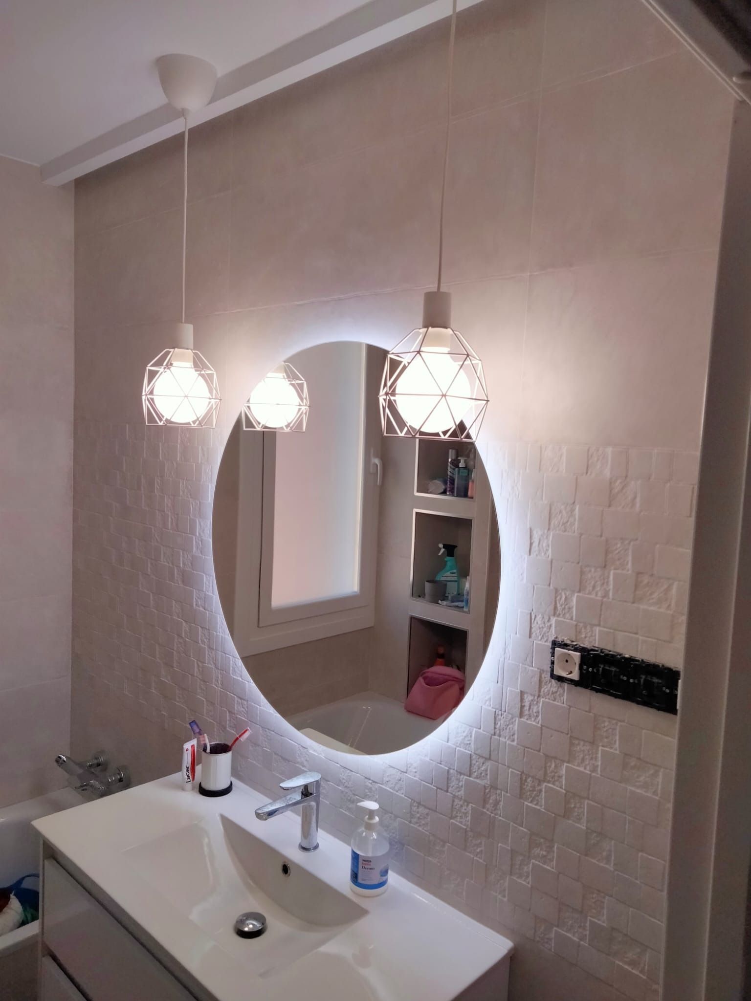 Baño con espejo ovalado retroiluminado, dos lámparas colgantes, lavabo blanco y azulejos decorativos de pared.