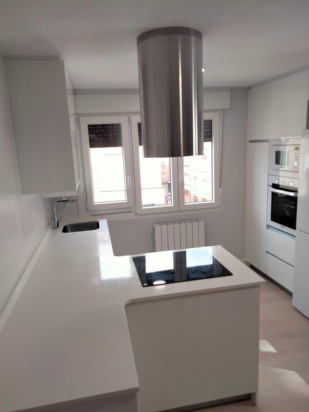 Cocina blanca moderna con isla, anafe y campana extractora de acero inoxidable frente a las ventanas.