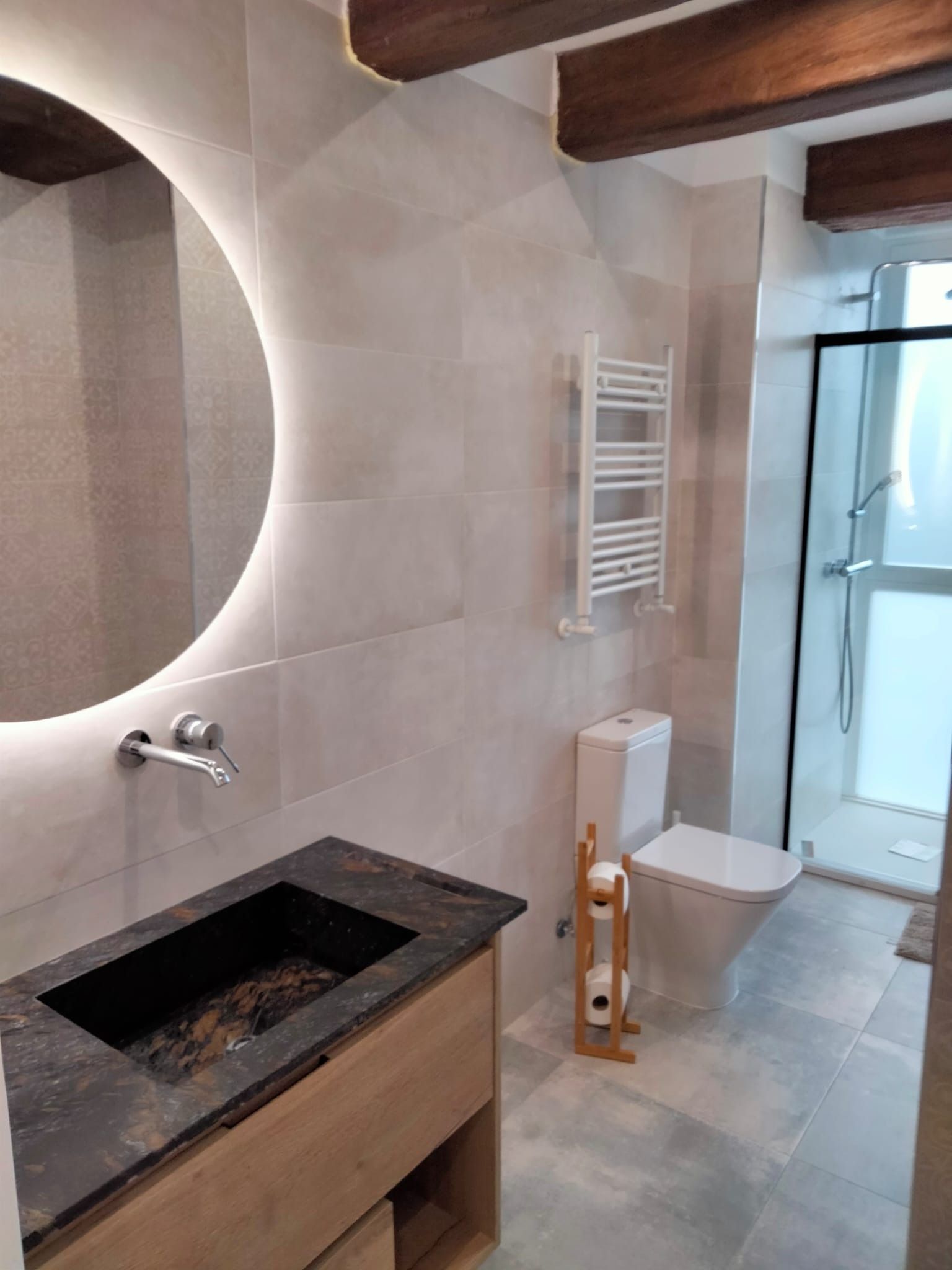 Baño moderno con azulejos gris claro, tocador de madera y un gran espejo redondo.