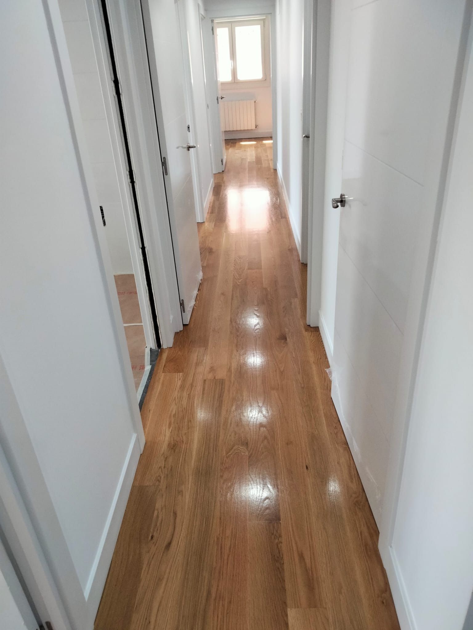 Pasillo estrecho con suelo de madera marrón claro, paredes y puertas blancas. Luz solar brillante al final.