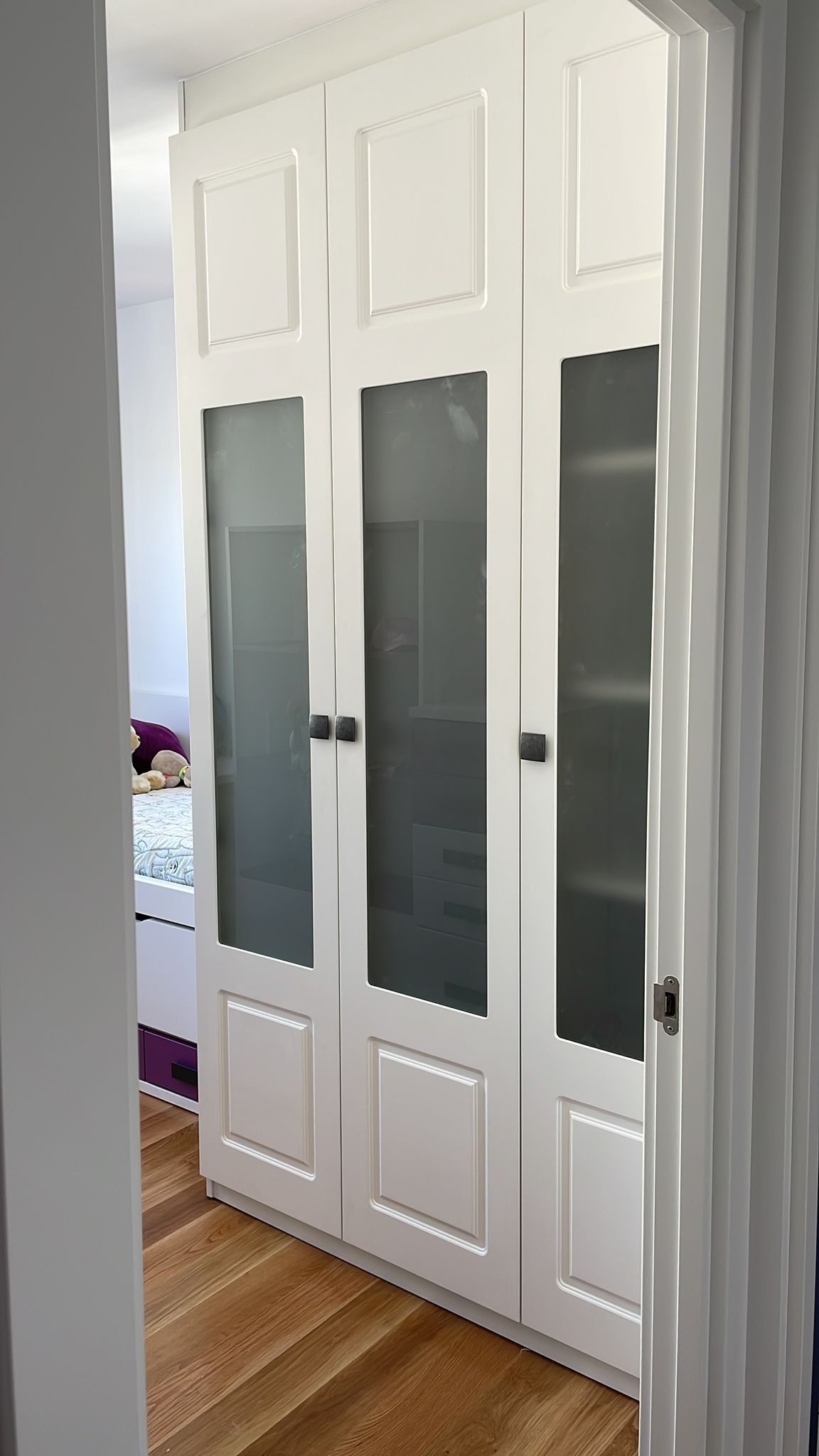 Armario blanco de tres puertas, paneles de vidrio esmerilado, manijas negras, ubicado en una habitación con piso de madera.