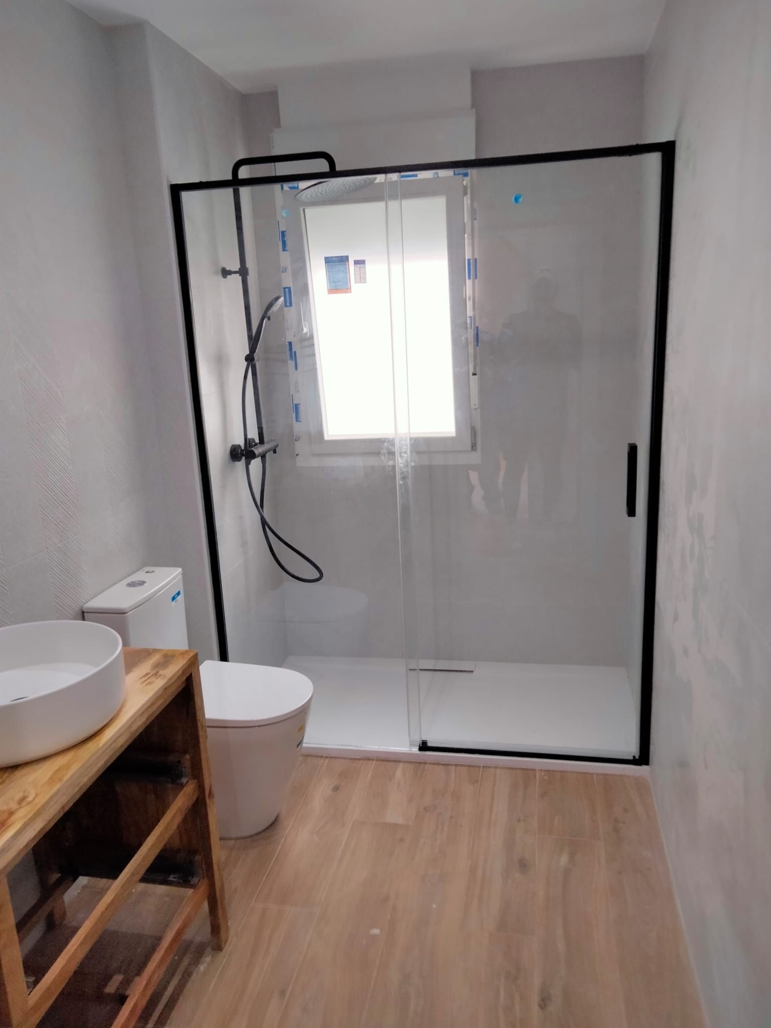 Baño con ducha a ras de suelo, inodoro y lavabo de madera. La ducha tiene marco negro. Suelo imitación madera clara.