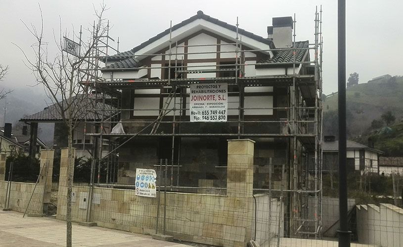 Casa en construcción con andamios, pared de piedra beige y ladera al fondo.