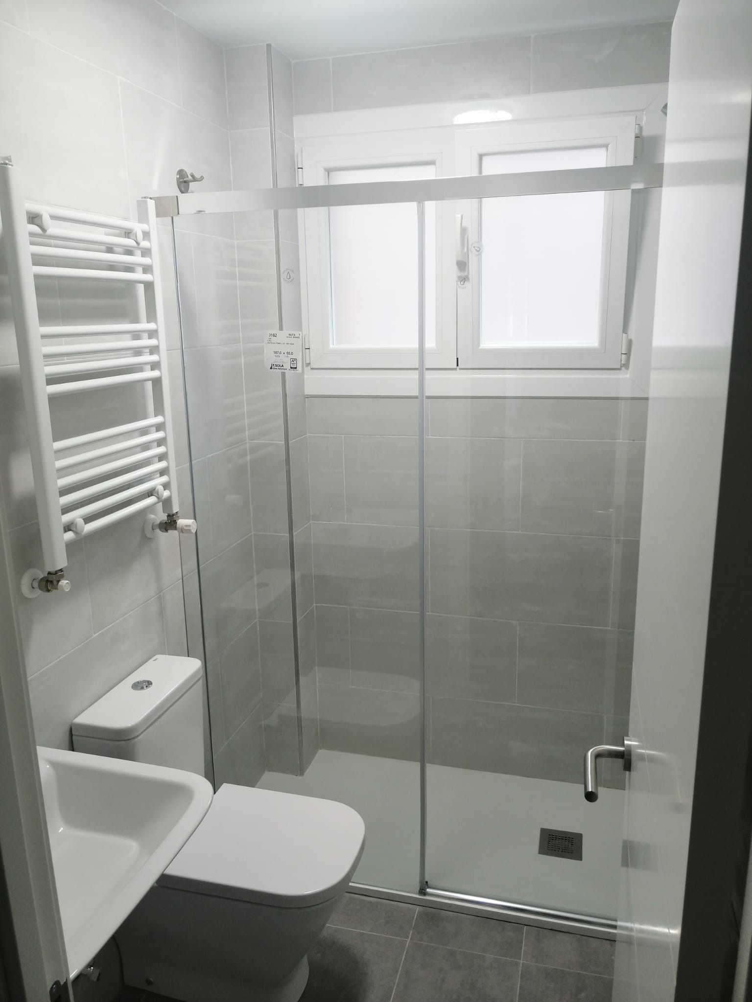 Baño pequeño y moderno con ducha de cristal, inodoro y toallero eléctrico. Paredes y suelo grises.