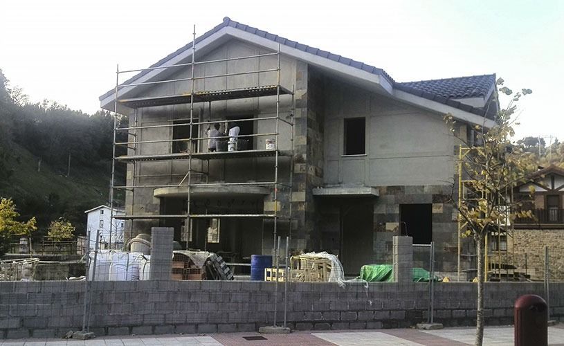 Casa en construcción con andamios y fachada de piedra; obreros presentes.