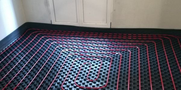 Sistema de calefacción por suelo radiante con tubos rojos colocados sobre una rejilla negra, cerca de una pared blanca.