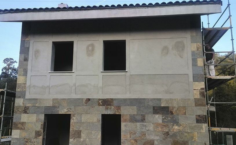 Fachada de edificio en construcción con base de piedra y nivel superior enlucido; andamios en el lateral.