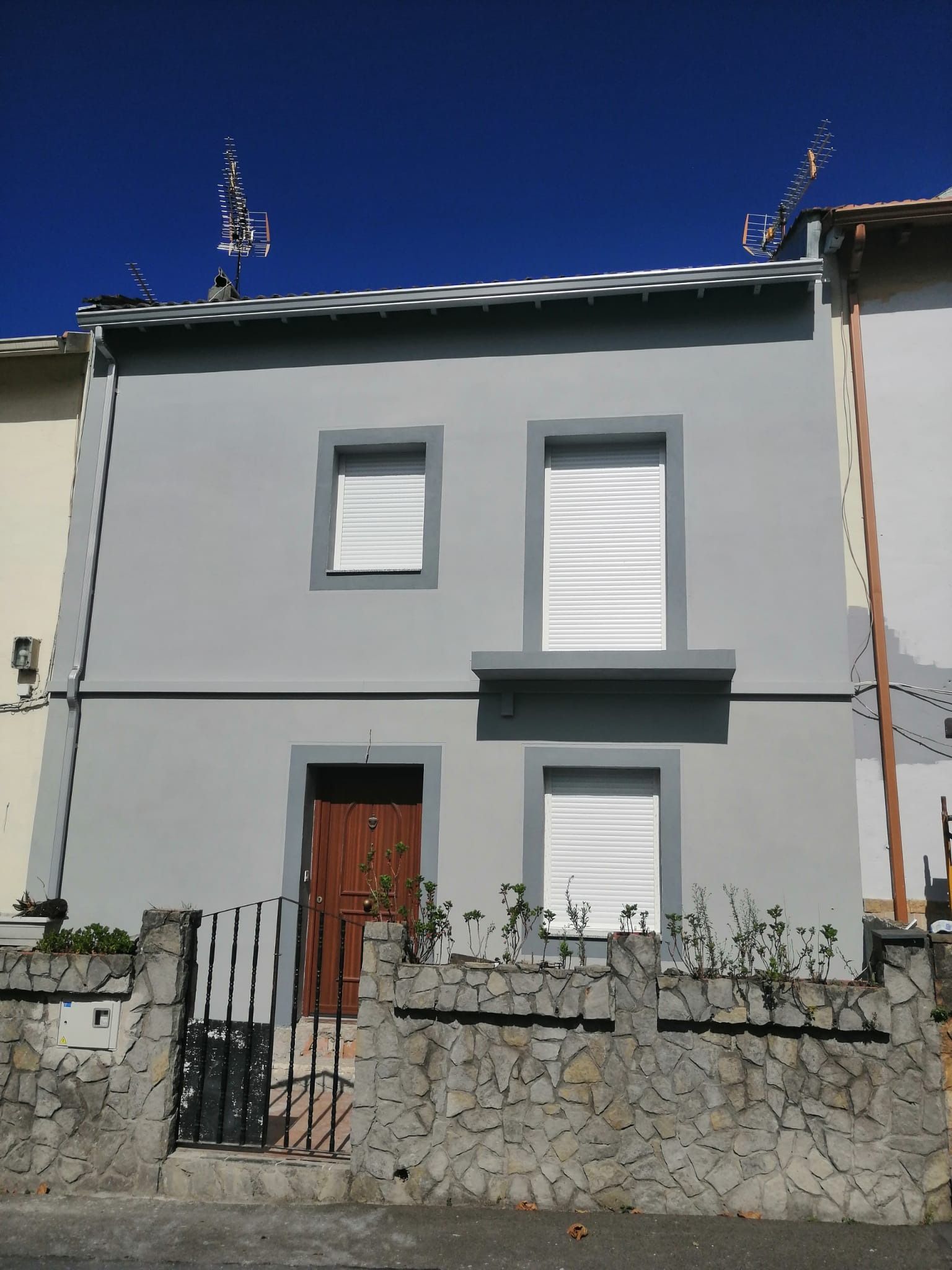 Casa gris de dos pisos con cortinas blancas, puerta marrón y pared de piedra en el frente.