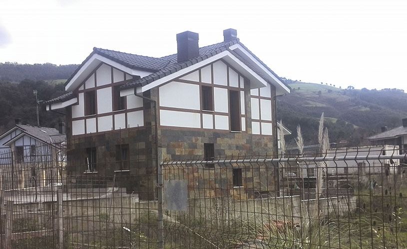 Casa de dos pisos con techo oscuro y nivel inferior de piedra, contra un fondo montañoso detrás de una cerca.