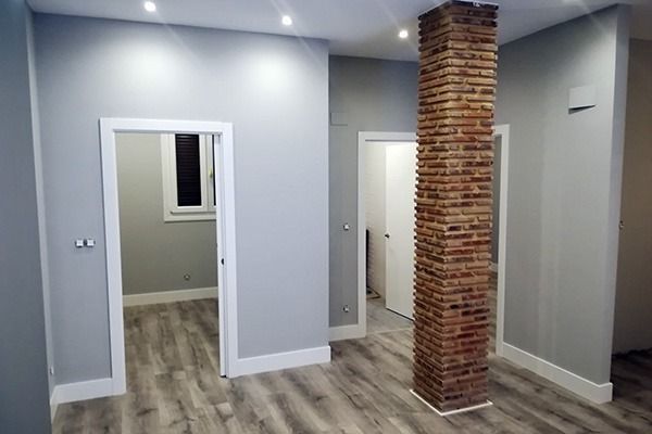 Vista interior de una habitación con paredes grises, suelo de madera y una columna de ladrillo. Se ven dos puertas.