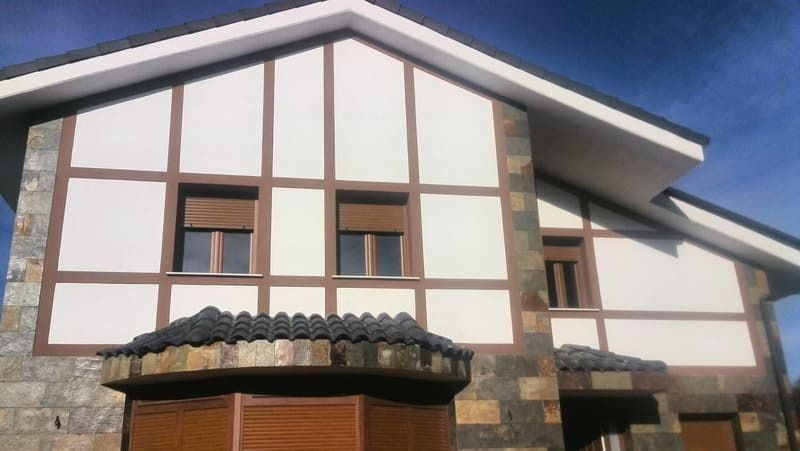 Casa con paneles blancos, vigas marrones y fachada de piedra; contraventanas marrones.