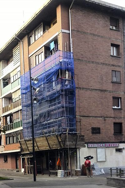 Edificio en construcción, con andamios cubiertos con malla azul. Una persona está de pie cerca.