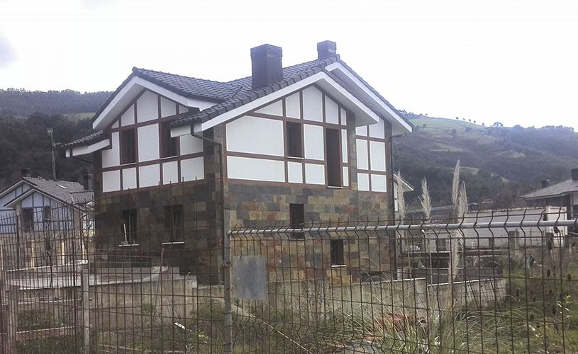 Casa de dos pisos con base de piedra, revestimiento blanco y marrón, techo oscuro y dos chimeneas, detrás de una cerca de alambre.