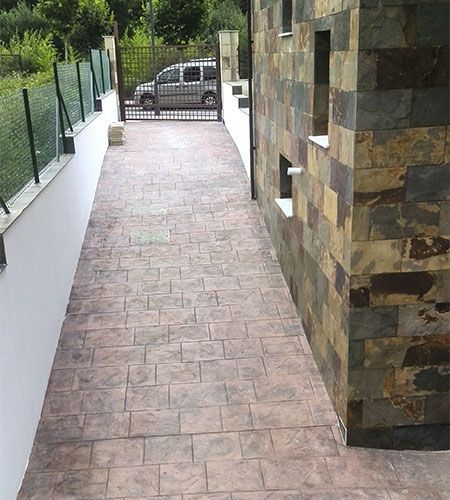 Largo camino con pavimento texturizado, junto a un muro de piedra y un muro blanco con valla. Puerta y vehículo al fondo.