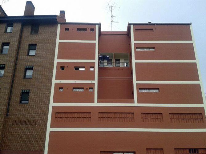 Fachada de edificio con molduras rojas y blancas, con pequeñas ventanas y un edificio de ladrillo adyacente.
