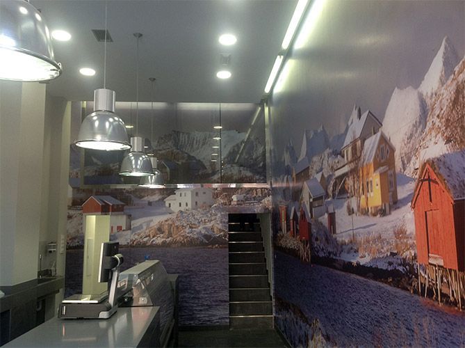 Interior de una tienda con un mural de montañas nevadas y un pequeño pueblo, mostrador de metal y luces.