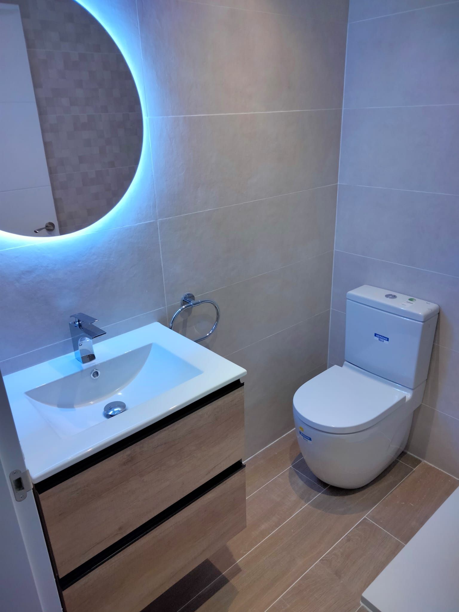 Baño moderno con espejo redondo, tocador e inodoro. Azulejos beige y mueble con efecto madera.