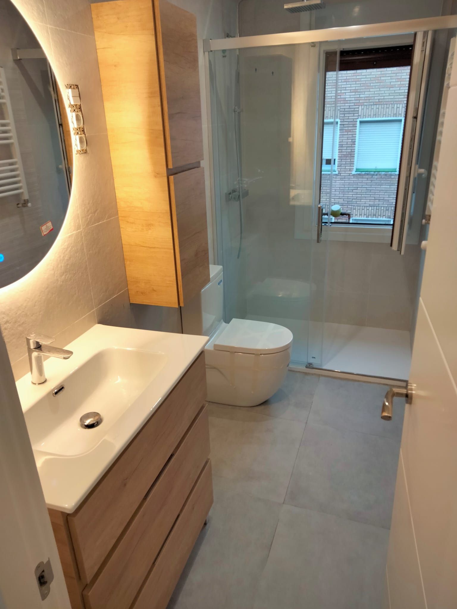 Baño moderno con tocador de madera clara, espejo redondo y ducha de cristal.