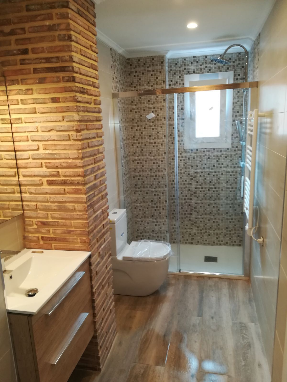 Baño con pared con acento de ladrillo, ducha de azulejos, piso con apariencia de madera y un inodoro.
