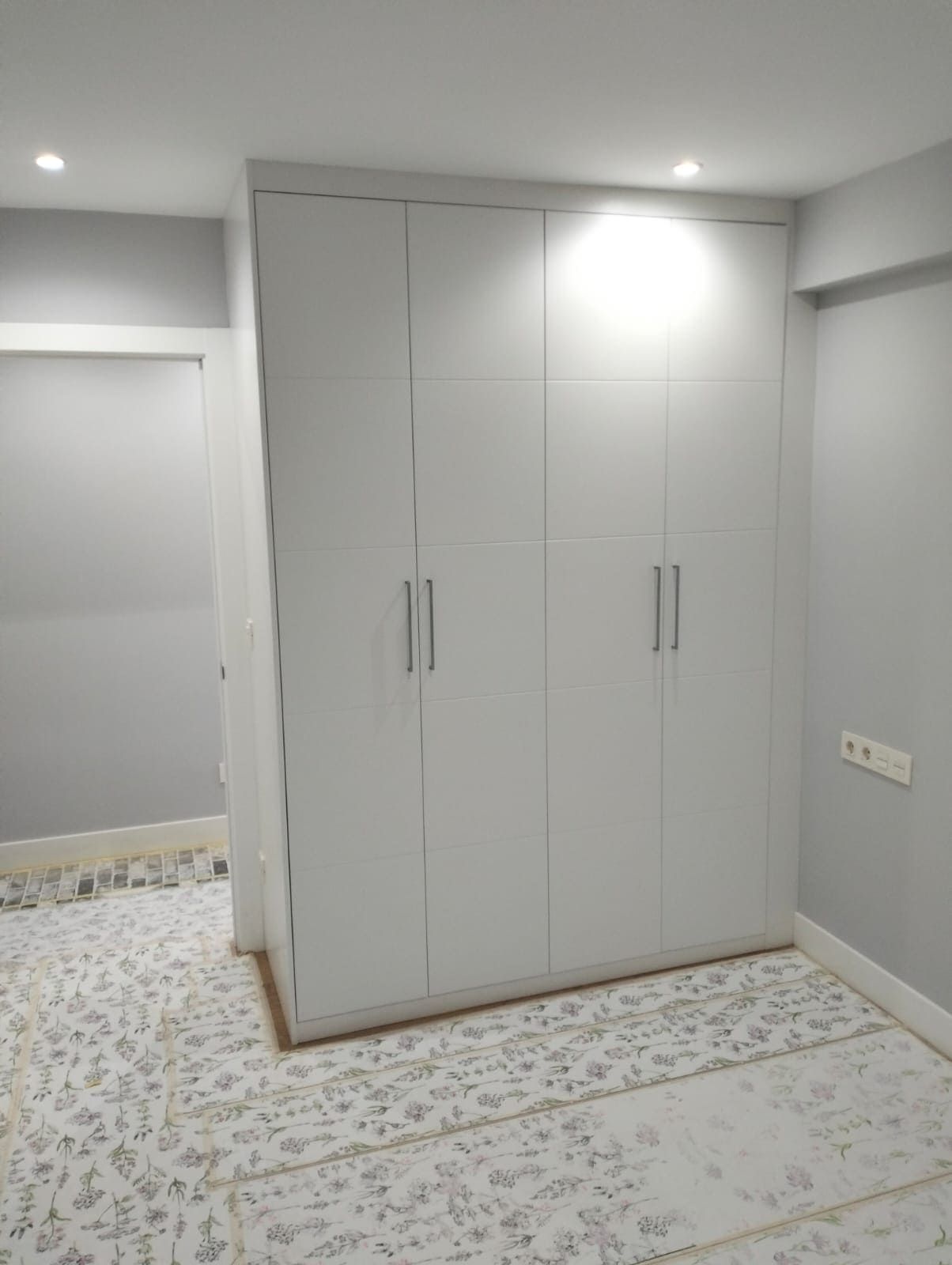 Armario empotrado blanco de cuatro puertas en una habitación con paredes gris claro y suelo estampado.