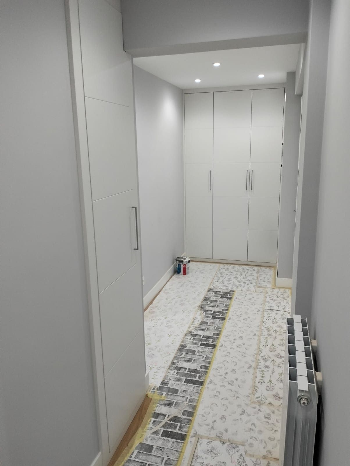 Pasillo blanco estrecho con armarios empotrados y radiador a la derecha. Suelo sin terminar, con azulejos decorativos.
