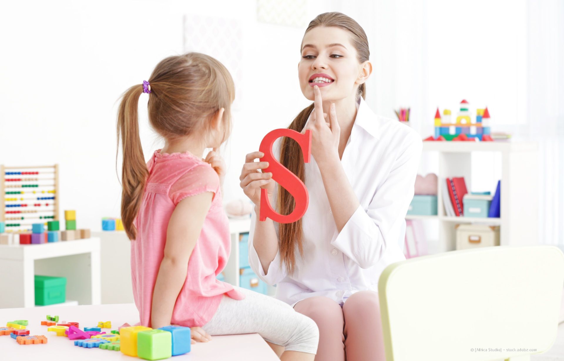 Frau und Kind spielen mit buntem Spielzeug in einem hellen Spielzimmer, während sich die Frau an einem Schminktisch Lippenstift aufträgt.