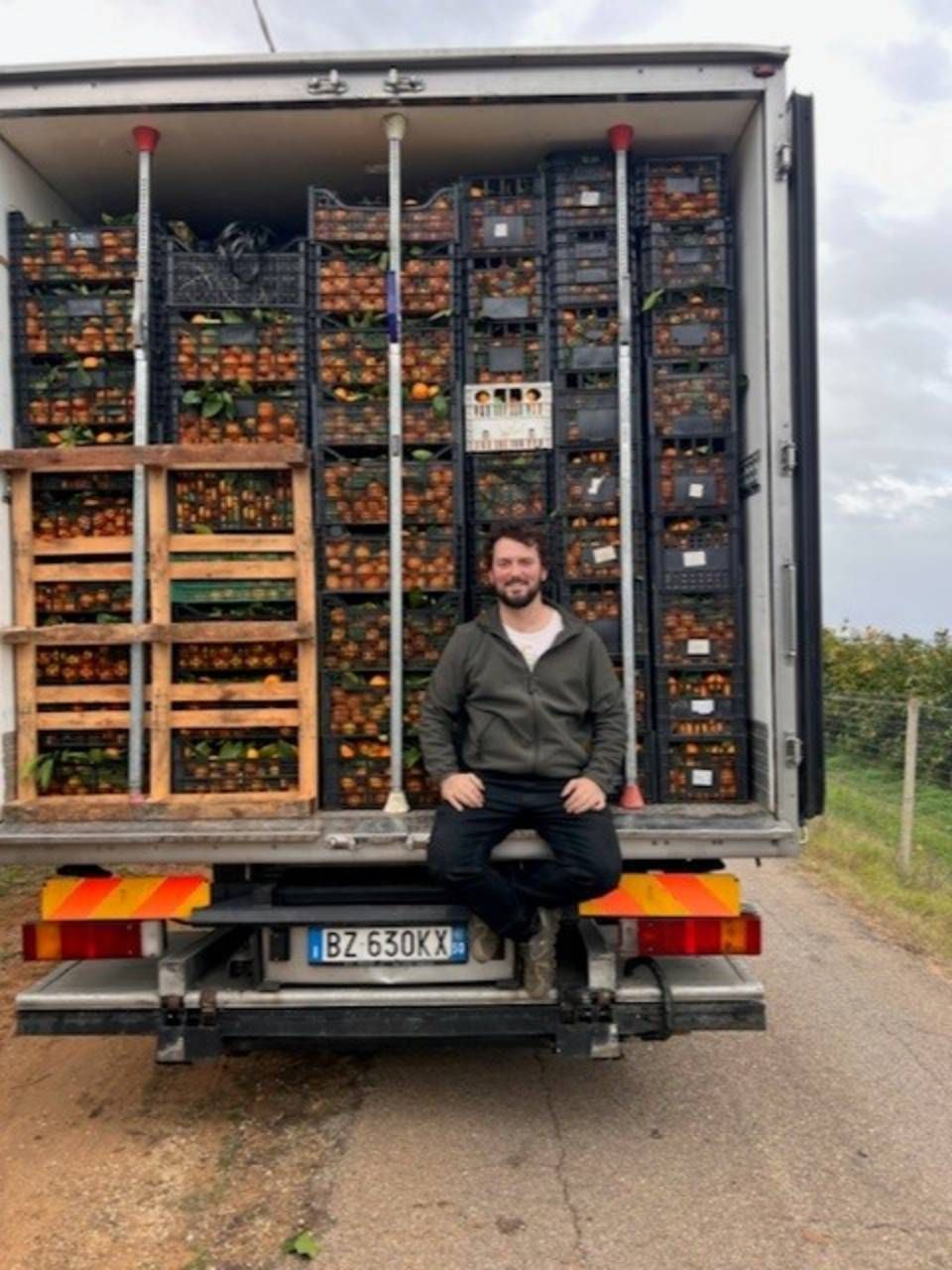 Uomo seduto sul portellone posteriore di un camion carico di casse di arance. Strada di campagna.