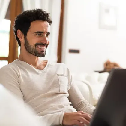 Un hombre con cabello oscuro y barba sonríe mientras trabaja con una computadora portátil en el interior.