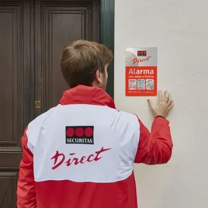 Una persona con una chaqueta roja y blanca de Securitas Direct instala un cartel de alarma en una pared cerca de una puerta.