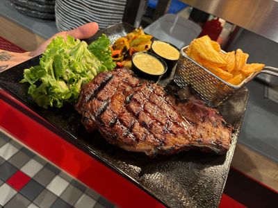 Steak grillé sur une assiette, accompagné de salade, de légumes et de frites, le tout sur une nappe à carreaux.