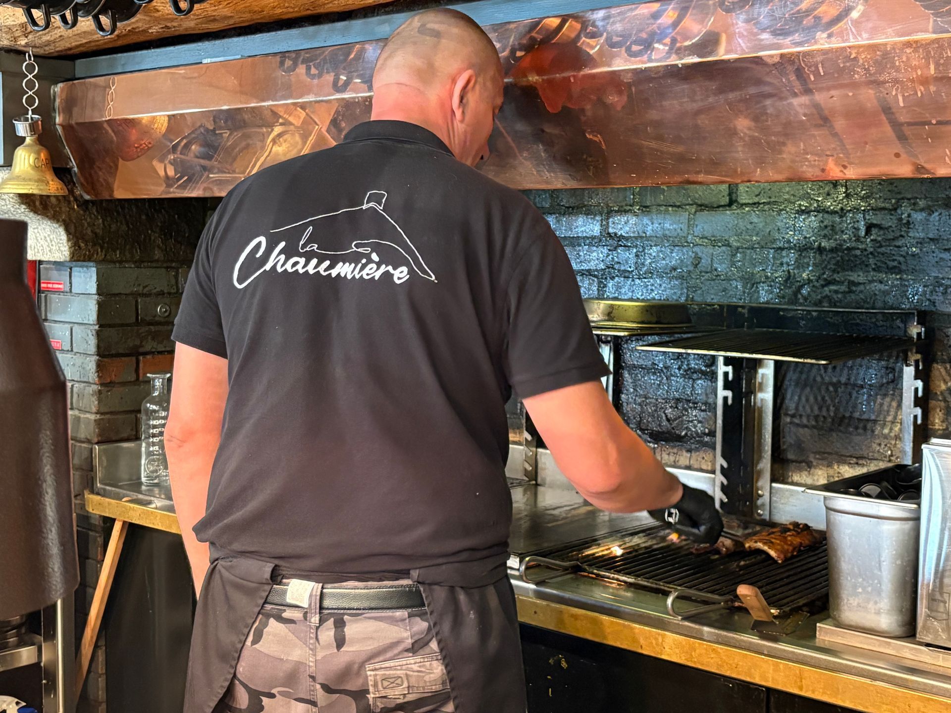 Une personne en chemise cuisine sur un gril dans une cuisine, avec une casserole et des ustensiles à proximité.