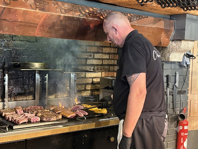 Un homme fait griller différentes viandes sur un barbecue sous une hotte en briques, d'où s'échappe de la fumée.