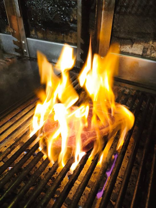 Des flammes s'élèvent d'une grille de barbecue à l'intérieur, près d'une cheminée vitrée ou de la porte d'un four.