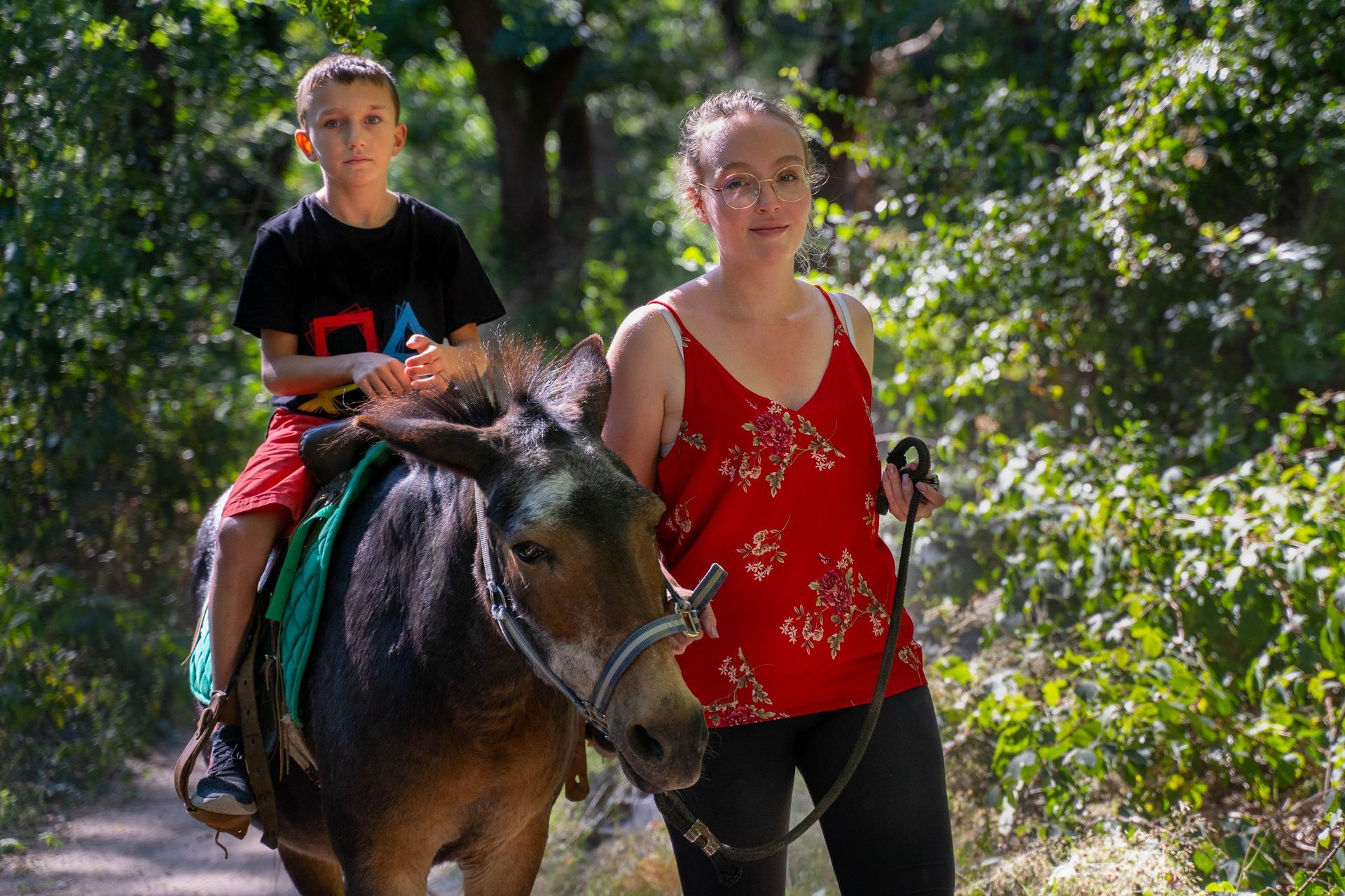 Un garçon à poney est guidé par une femme sur un sentier. Journée ensoleillée, décor forestier.