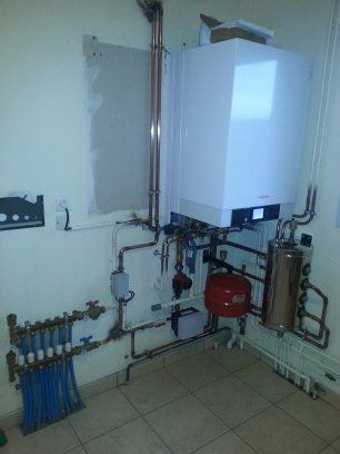 Une chaudière à gaz murale avec tuyaux en cuivre, vase d'expansion rouge et collecteur de chauffage au sol dans une pièce